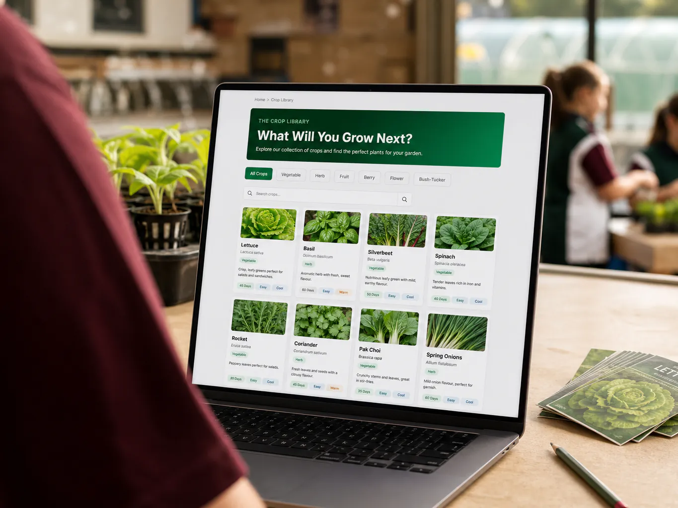 A teacher's laptop in a classroom showing the Crop Library, a searchable grid of crop profiles with hydroponic yield and water-saving data.