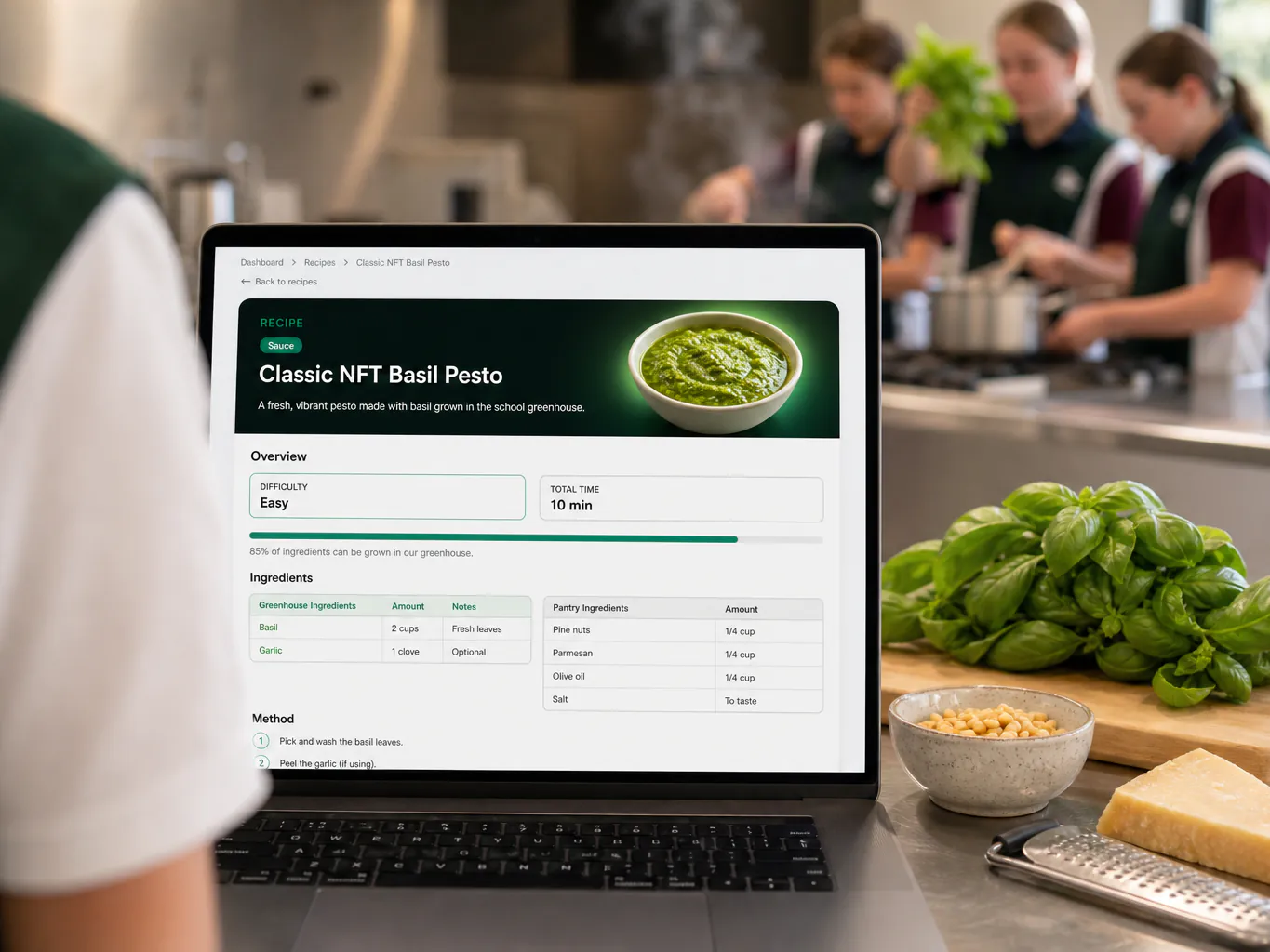 A teacher's laptop in a school kitchen showing the Recipe Book open on the Classic NFT Basil Pesto recipe, with ingredients and method.
